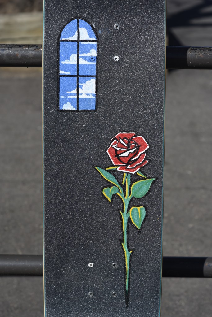 A griptape painting depicting a simple red rose and an arched window with a blue sky and clouds beyond it